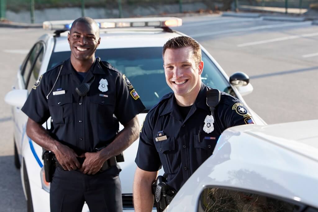 Two smiling responsible, honorable policemen serving on the job with police cars in the foreground and background.  Their contribution makes them admirable.