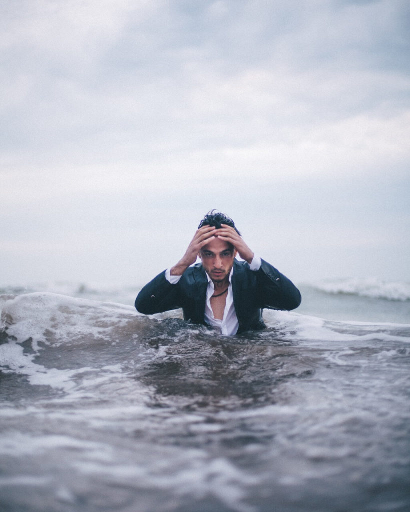 Wonderiing about the way we dress, we see a  perplexed young man stoically pushing through high water and waves while wearing a blue business suit.