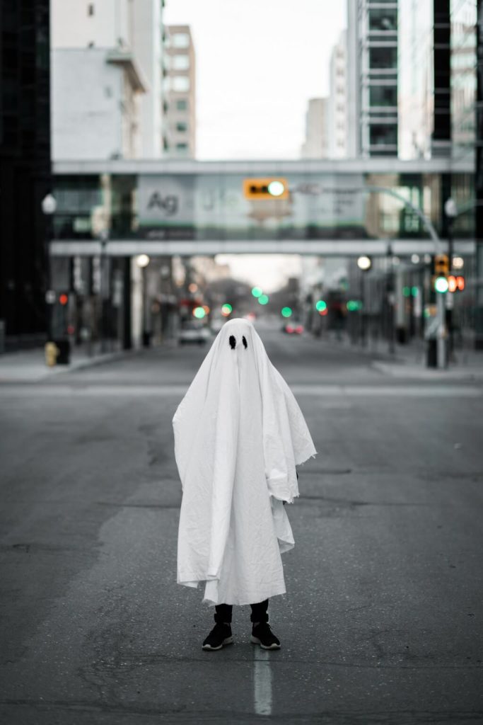 In a ghost costume, a man stands on a city street representing what we are afraid of. The bottom of his pants and his shoes remain showing.