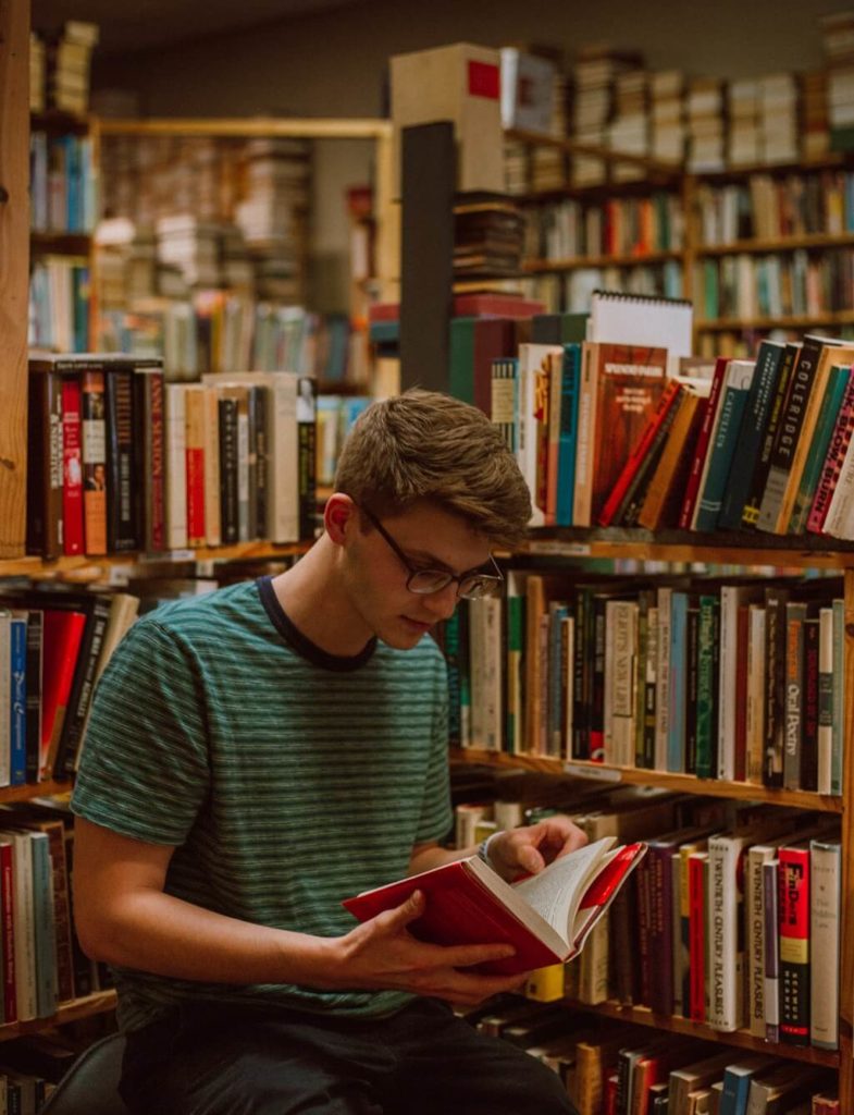 The photo shows a young man with a deep interest in a book as he is surrounded by shelves of books.