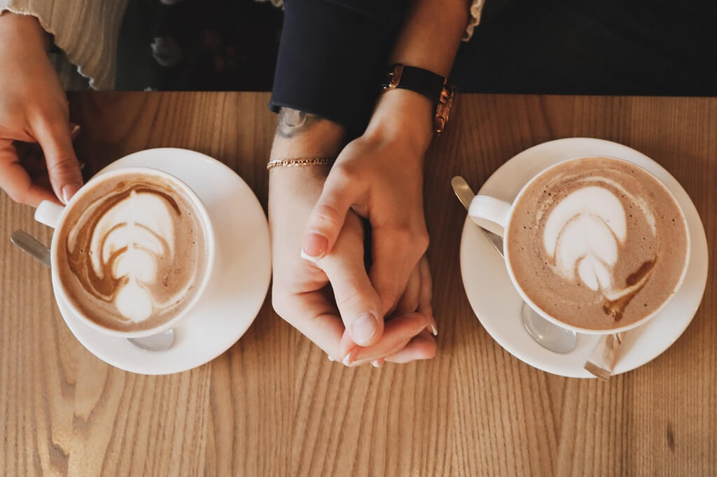 How to mingle is shown in this photo. The hands of two people are holding each other, 
resting on a table as they have coffee together.