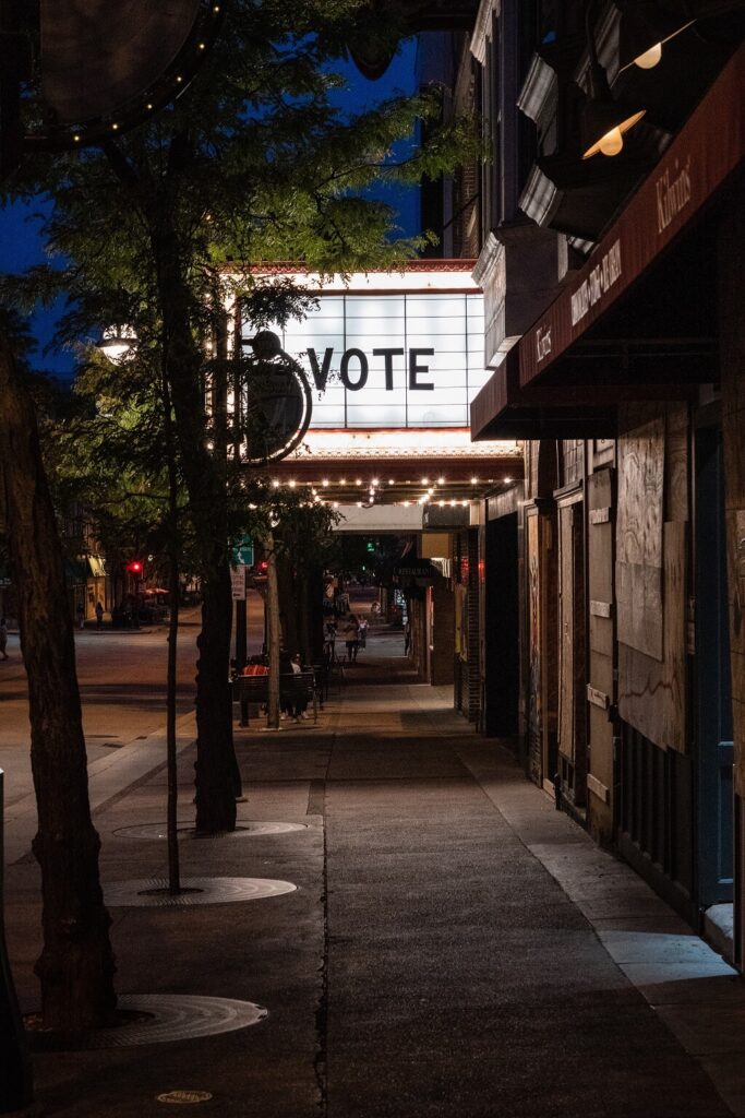 Reminding us of our promises, the photo shows a city sidewalk in the evening approaching the lit-up marquee of a movie theater that has the large word VOTE written on it.